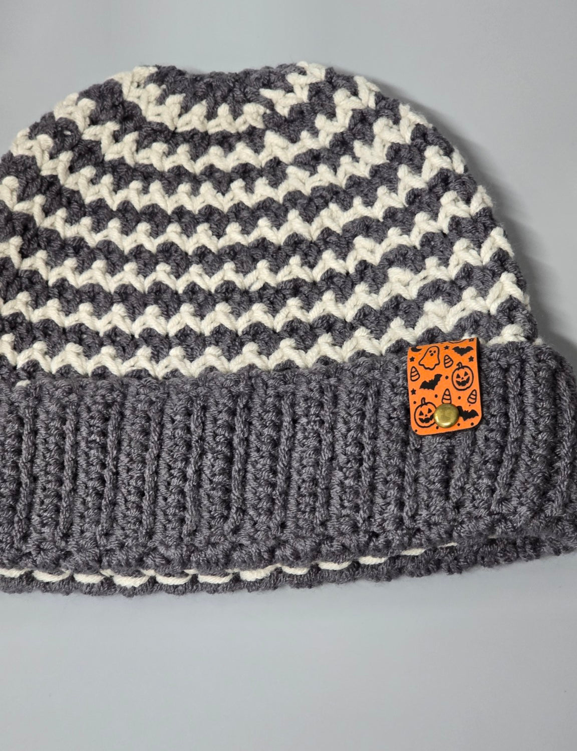 Orange Halloween fold-over tag attached to the brim of a gray and cream crochet beanie. Beanie shown as a display example.